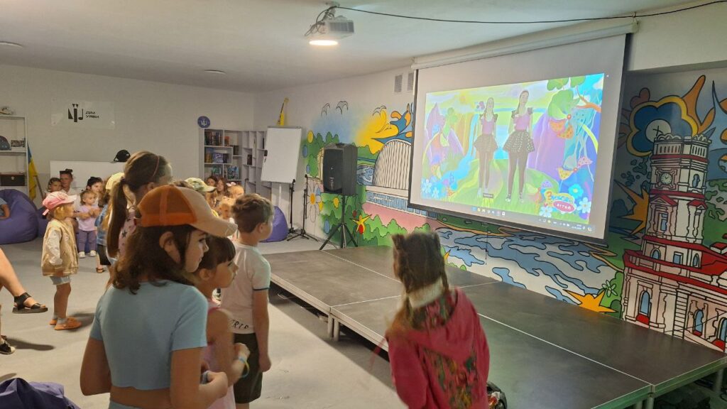 В укритті. оснащеному мультимедійним обладнанням (проєктор, екран) діти різного віку стоять на майданчику перед великим екраном, на якому видно двох дівчат на фоні фантастичного різнобарвного пейзажу.